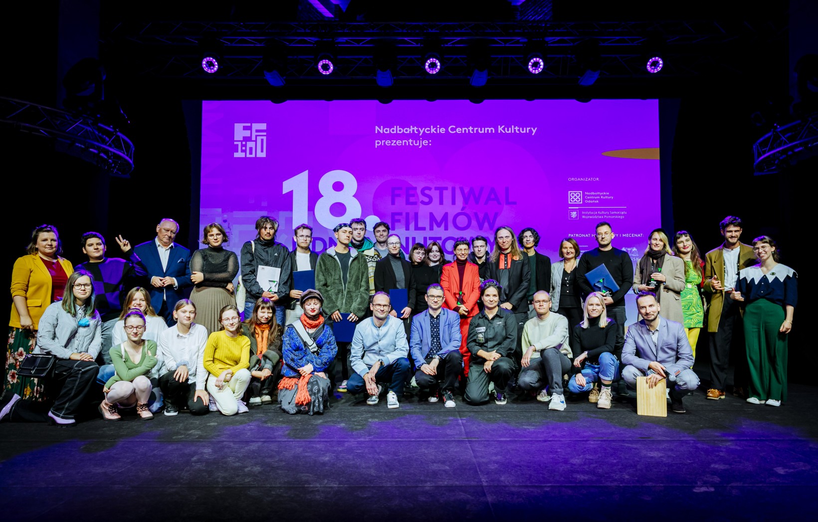 zdjęcie grupy ludzi na scenie, trzymają dyplomy, w głębi ekran, napis "18 festiwal filmów jednominutowch