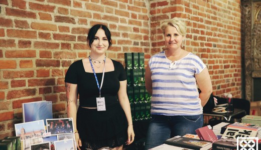 zdjęcie dwóch kobiet na tle ceglanej gotyckie ściany Centrum św. Jana. Kobiety stoją za sklepową ladą, na której leżą publikacje i gadżety