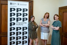 zdjęcie wewnątrz, trzy kobiety uśmiechają się do obiektywu. Stoją przy roll-upie z napisem Pracownia Kultury i logotypem NCK