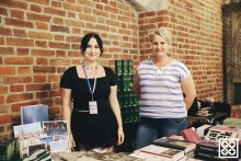 zdjęcie dwóch kobiet na tle ceglanej gotyckie ściany Centrum św. Jana. Kobiety stoją za sklepową ladą, na której leżą publikacje i gadżety