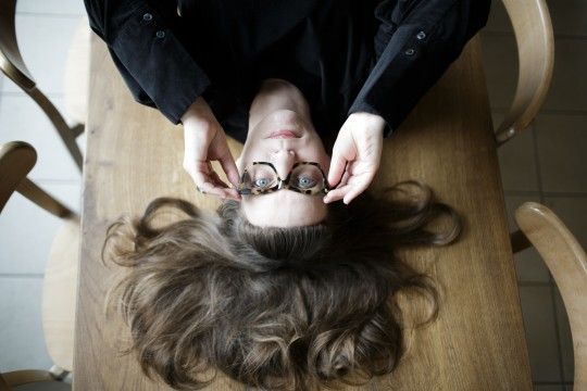 Zdjęcie przedstawia reporterkę Magdalenę Grzebałkowską. Zdjęcie jest kadrowane z góry. Kobieta leży na stole i dłońmi podtrzymuje oprawki okularów, które ma na twarzy. Stół jest drewniany. Wokół niego stoją drewniane krzesła. Kobieta ma długie włosy.