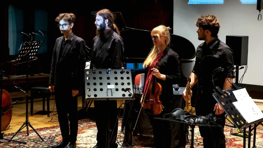 Czworo artystów w trakcie oklasków. Dwoje z muzyków trzyma w dłoniach instrumenty akustyczne. W tle fortepian.