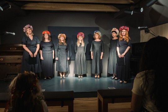 Siedem kobiet w czarnych sukniach. Stoją na scenie, na głowie mają wianki.