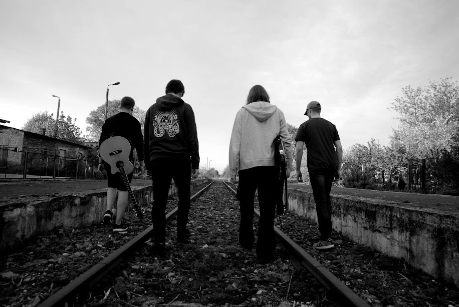 Czterech muzyków z gitarami, odwróconych tyłem, idą po torach wzdłuż peronu, zdjęcie czarnobiałe