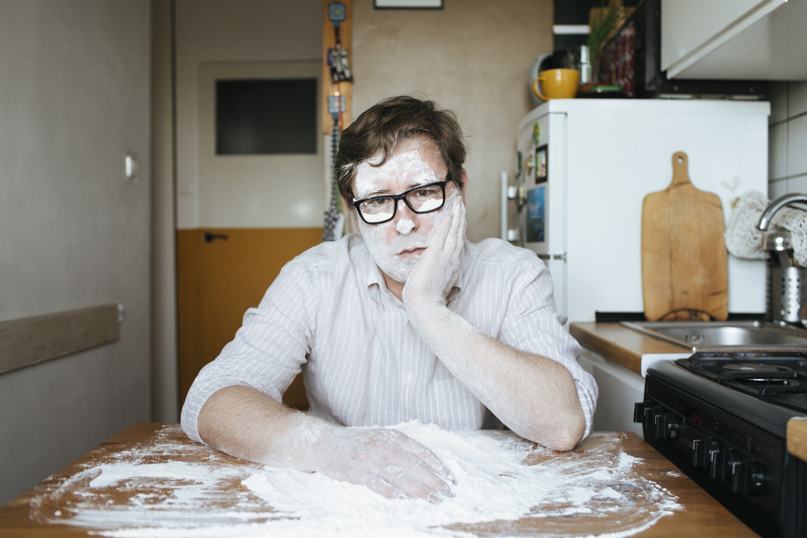 Zdjęcie poety Tadeusza Dąbrowskiego siedzącego w kuchni. Mężczyzna nosi okulary w grubych oprawkach i prawą rękę w mące. Lewą ręką podtrzymuje twarz umorusaną w mące tak jak ręce.