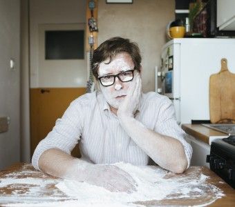 Zdjęcie poety Tadeusza Dąbrowskiego siedzącego w kuchni. Mężczyzna nosi okulary w grubych oprawkach i prawą rękę w mące. Lewą ręką podtrzymuje twarz umorusaną w mące tak jak ręce.