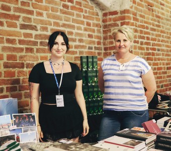 zdjęcie dwóch kobiet na tle ceglanej gotyckie ściany Centrum św. Jana. Kobiety stoją za sklepową ladą, na której leżą publikacje i gadżety