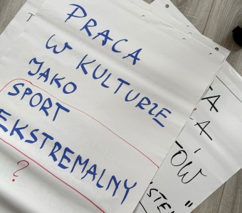 na drewnianej podłodze leży kartka z napisem "praca w kulturze jako sport ekstramalny?"
